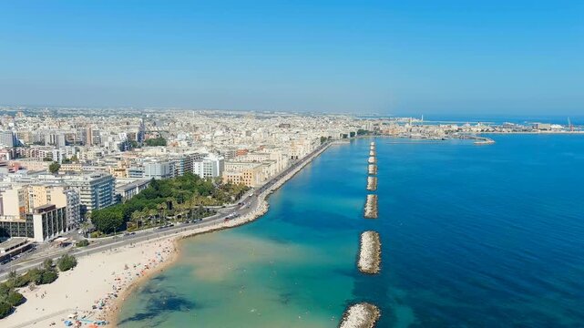Bari, Italy. The central Lungomare embankment, Drone footage over the Adriatic with stone breakwaters, sandy beach, and dense white cityscape under clear summer skies. Drone footage, Departure of the