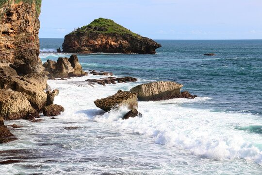 Beautiful tropical coast with rolling white waves crashing against rugged rocks and an island in the background.