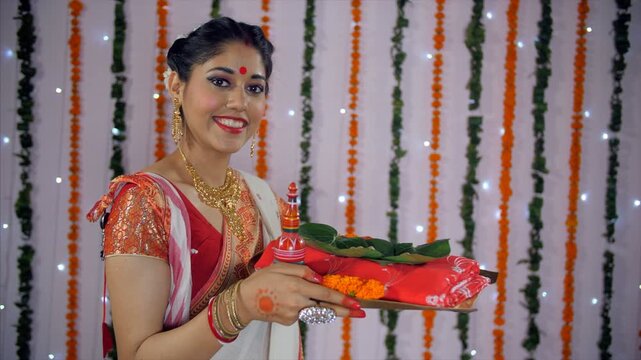 Bengali woman in ethnic saree happily holding a plate with religious offerings. Pretty Indian female smiling while holding a Durga Puja thali with sindoor  red saree  betel leaf (Paan Patta)  and s...
