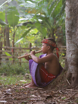 An Indonesian village boy in traditional clothes sitting under a tree and playing a bamboo flute in a peaceful rural setting.
