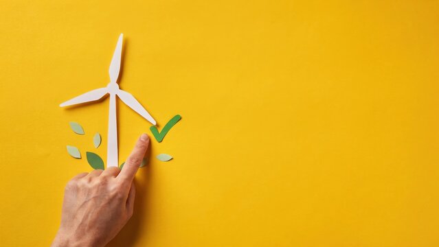 A hand holds a wooden wind turbine model with green leaves on a yellow background.