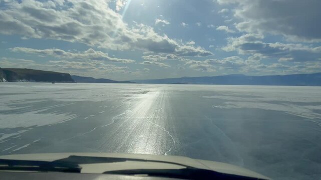 POV view from a vehicle driving on the frozen surface of Lake Baikal with sun glare, textured ice and distant mountains under a cloudy sky in Russia