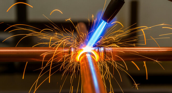 Copper pipe brazing with a gas blowtorch. Intense blue flame heating a metal joint creating bright orange sparks. Professional plumbing and HVAC repair concept for industrial maintenance marketing.
