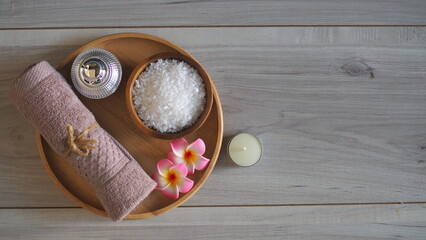 Luxury spa still life with white bath salt in a wooden bowl, a soft pink mauve towel tied with jute rope, a metallic cosmetic jar, pink frangipani flowers, and a candle on a light gray wooden table. P © kanjana