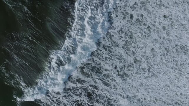 Aerial top down footage of powerful ocean surf rolling in and breaking into white sea foam near the shoreline, creating a dynamic natural water texture background