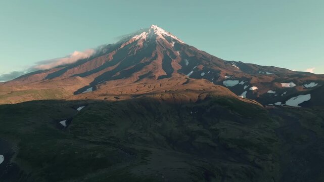 Aerial footage of Koryaksky Volcano with snow patches and clouds in warm sunrise light, rugged volcanic slopes and tundra hills in Kamchatka Krai, Russia