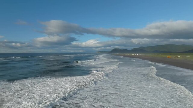 Drone view of surf rolling onto black volcanic sand at Khalaktyrsky Beach near Petropavlovsk-Kamchatsky, Kamchatka Krai, Russia, with mountains and clouds
