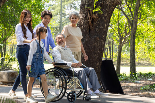 Happy multi-generational Asian family taking a walk in a sunny park with elderly grandfather in a wheelchair, enjoying quality time, healthcare, and outdoor togetherness