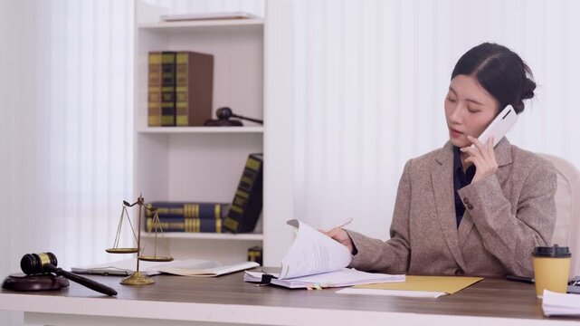 Asian female lawyer attorney talking on phone reviewing legal documents case files in law firm office with gavel justice scales background consulting client
