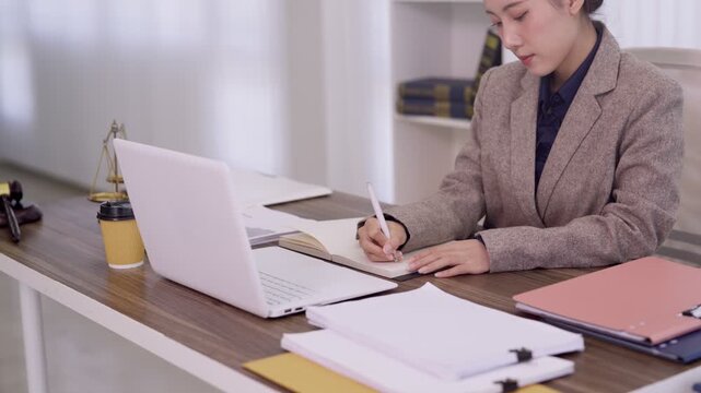 Asian lawyer woman reading legal documents writing notes using laptop computer working at office desk with justice scales studying case files professional