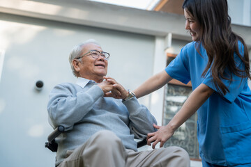 Fototapeta premium Elderly man sits in a wheelchair outside a building. Woman in blue scrubs assists him. He smiles as she helps. Professional caregiver supports senior well-being