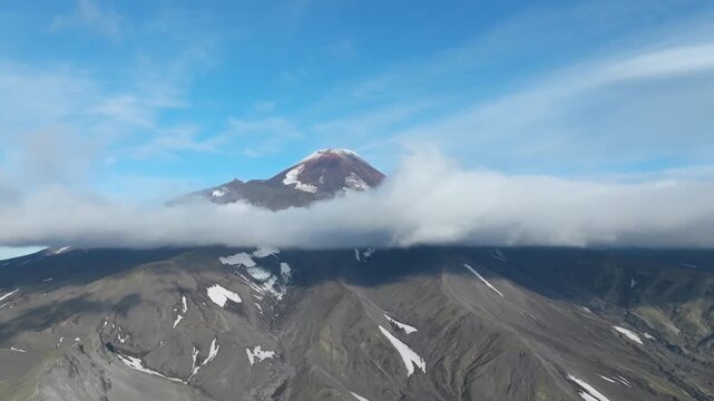 Aerial footage of Avachinsky Volcano with a cloud layer wrapping the slopes, rugged volcanic landscape and snow patches under blue sky, Kamchatka Krai, Russia