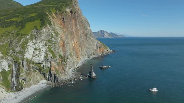 Drone footage of rugged sea cliffs, green slopes and a small beach on Avacha Bay, Kamchatka Krai, Russia, with calm Pacific water and a boat offshore