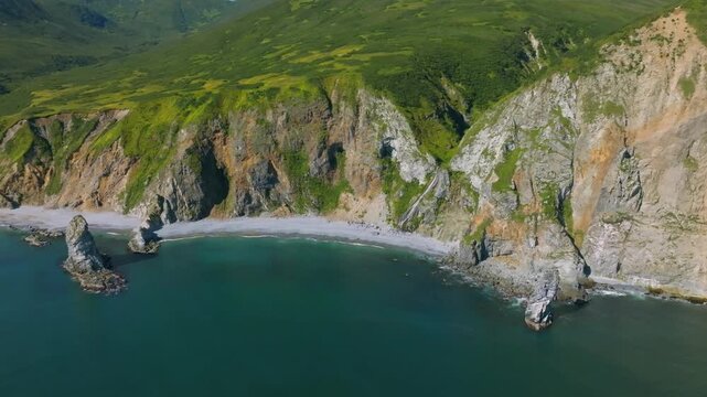 Drone footage of rugged sea cliffs, green slopes and a narrow pebble beach along Avacha Bay on the Kamchatka Peninsula, Russia, with calm ocean water and summer light