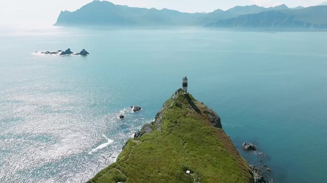 Aerial footage of a solitary lighthouse on a green rocky cape above Vilyuchinskaya Bay, Kamchatka, Russia, with calm blue sea, cliffs and distant mountains
