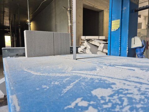 Low angle worm's-eye view of a band saw table and blade with an AAC concrete block and debris in the background.