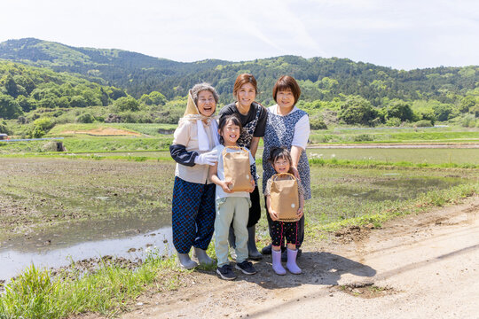 里山の水田で収穫物を手に笑顔を見せる三世代の日本人女性と子供