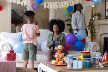 Obraz premium African American family celebrating child birthday in living room, holding cake and fixing balloons