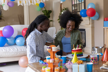 Obraz premium African American couple celebrating birthday in living room, blowing candles on rainbow cake