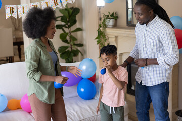 Obraz premium African American family celebrating in living room with child blowing blue balloon near white sofa