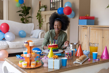 Obraz premium African American woman in green shirt and headband sitting at home blowing candles on rainbow cake