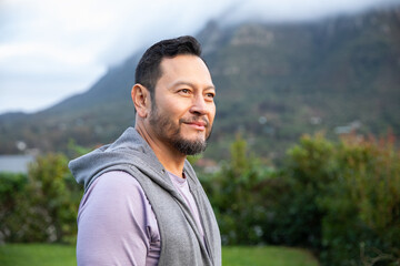 Mature Asian man standing outdoors near shrubs and mountains, wearing grey hoodie over purple shirt © wavebreak3