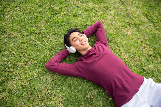 Asian man lying on grassy lawn wearing white headphones and maroon shirt and light gray sweatpants