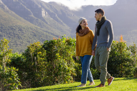 Diverse couple walking in park by low hedge and mountains, wearing beanie and scarves, copy space