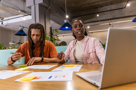 African American coworkers reviewing charts, sticky notes near laptop at office in orange and pink