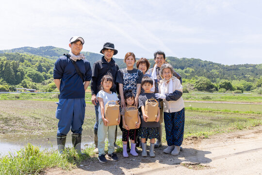 田植え後の水田で収穫物を手に笑顔を見せる三世代家族