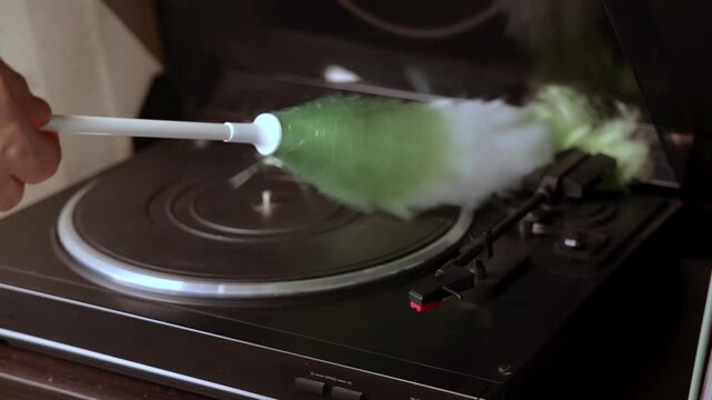 Hand holding a feather duster cleans a vintage turntable, showcasing the meticulous care of vinyl records in a cozy indoor setting with soft lighting
