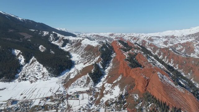 Drone footage of the Jeti Oguz gorge and the Seven Bulls red sandstone cliffs with snow-covered ridges, pine forest slopes and a valley settlement, Kyrgyzstan