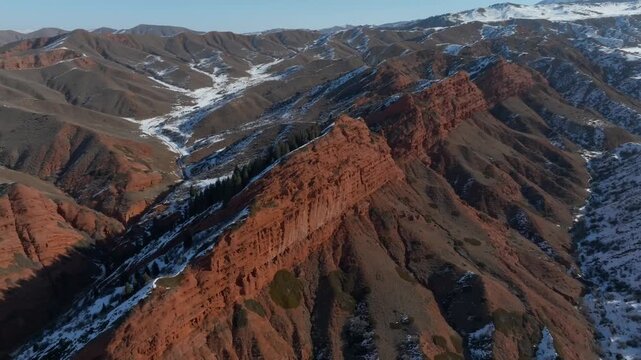 Drone footage of the Seven Bulls red sandstone cliffs in Jeti-Oguz Gorge, Issyk-Kul Region, Kyrgyzstan, with snowy mountain slopes and a winding valley