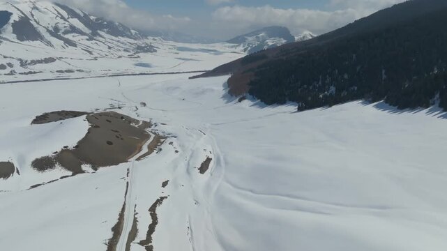 Wide aerial view of snow-covered mountains and a winding dirt road across a winter valley in Grigoriev Gorge, Issyk-Kul Region, Kyrgyzstan