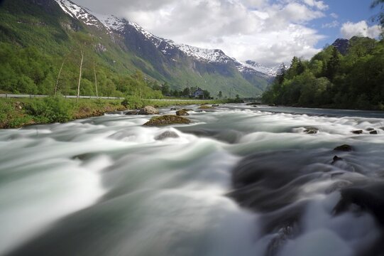 A wide river in a mountainous landscape with snow-capped peaks and cloudy skies, Olden, Nordfjord, Norway