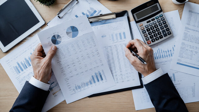 Businessman analyzing financial report, holding paper with charts, calculator and documents desk