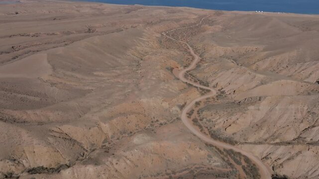 Drone footage of Skazka Canyon arid hills and winding dirt road with sparse shrubs, overlooking the blue waters of Issyk Kul Lake in Kyrgyzstan