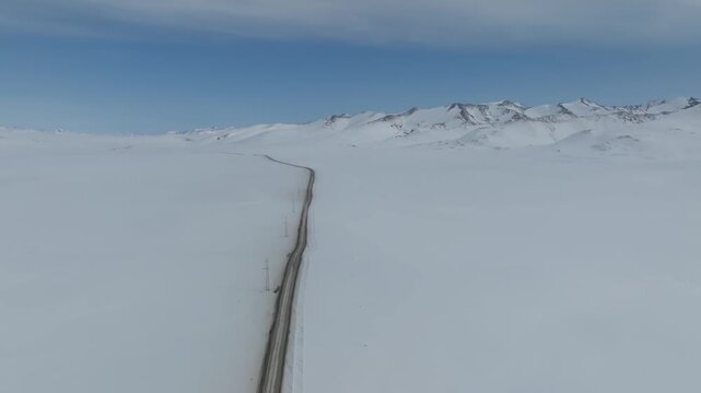 Drone footage of a long road cutting through the snow-covered Arabel Plateau, with distant mountain ridge under clear blue sky, Kyrgyzstan, Central Asia