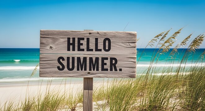 A wooden sign with the text hello summer on a sandy beach with ocean waves and tall grass