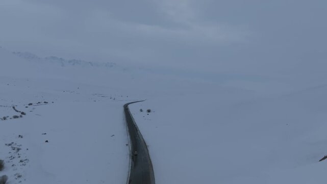 Drone view of a long asphalt road cutting through a vast snowy plateau at Ala-Bel Pass, Naryn Region, Kyrgyzstan, overcast winter weather and solitude