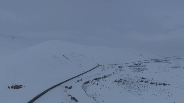Drone view of a winding highway crossing Ala-Bel Pass in the Tien Shan mountains, Kyrgyzstan, with snow-covered slopes, low clouds and remote winter landscape