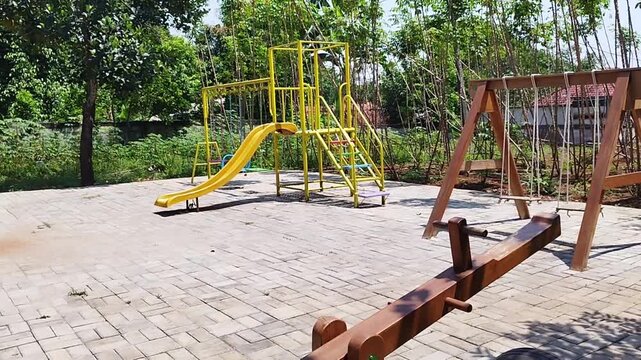 Panning shot of an empty outdoor children playground with a yellow slide, wooden swings, and seesaw surrounded by green trees