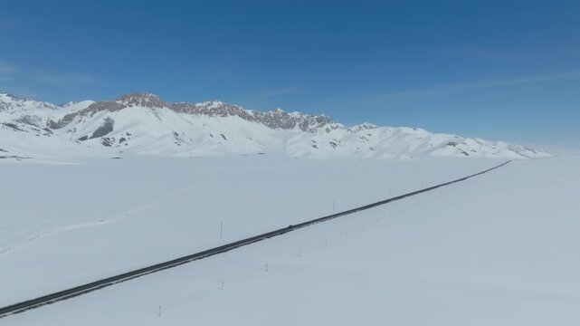 Aerial footage of a long highway cutting through the snow-covered Alay Valley with rugged mountains in the background, Osh Region, Kyrgyzstan on a clear day