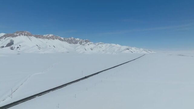 Drone footage of a long road crossing a vast snow-covered plain with rugged mountain range under clear blue sky in Alay Valley, Osh Region, Kyrgyzstan