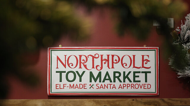 Festive Christmas holiday sign for north pole Toy Market featuring green and red font on a white background with decorative text Elf-Made Santa Approved hanging in a cozy Xmas shop display