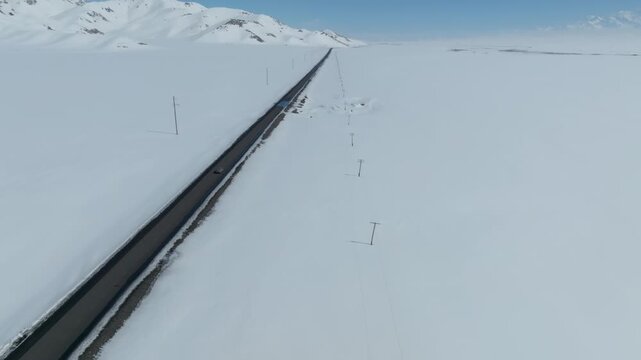 Drone footage of a long asphalt highway cutting through a vast snow-covered plain with mountain ridges in the distance, Alay Valley, Osh Region, Kyrgyzstan