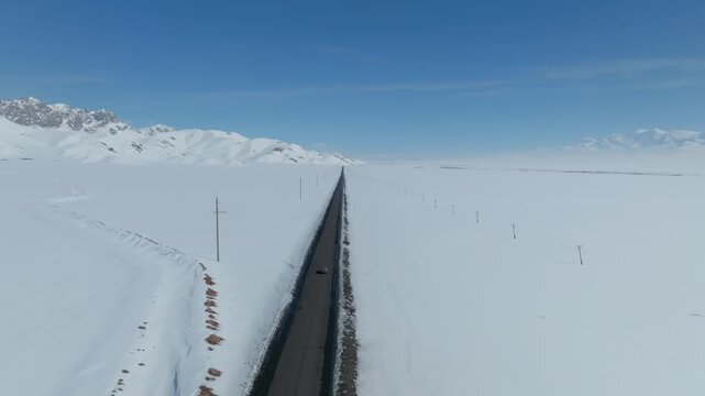 Drone footage of a long straight highway cutting through a vast snow-covered plain with distant mountains under blue sky in Alay Valley, Osh Region, Kyrgyzstan