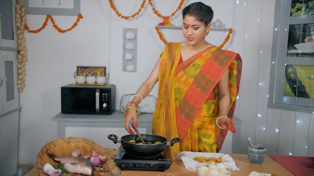 A Bengali wife grinding spices in a traditional grinder - Indian Food. An Indian lady in traditional Bengali saree using mortar and pestle to grind herbs while frying pakoras in her kitchen