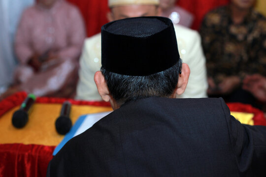 An over-the-shoulder shot of a man wearing a traditional Indonesian black velvet cap, known as a peci or songkok, and a dark suit jacket.