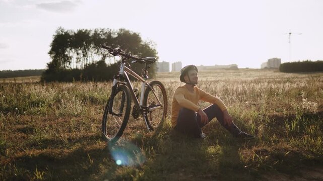Inspired man in helmet sitting near bicycle during break of cycling ride outdoor. Young man enjoying solo journey and with light smile looking around in sunny field at sunset.